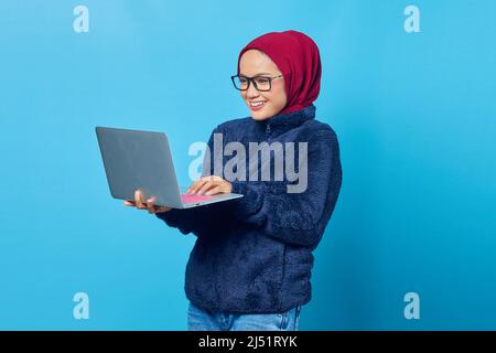 Schöne asiatische muslimische Student trägt blaue Jacke und Sonnenbrille lächelnd und hält Laptop Stockfoto