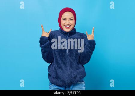 Porträt einer jungen asiatischen Frau in blauer Jacke, die Rock-Symbol mit Händen auf blauem Hintergrund macht Stockfoto