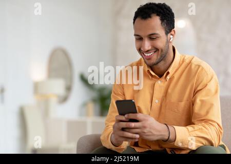 Glücklicher arabischer Mann, der Mobiltelefon mit kabellosen Kopfhörern benutzt Stockfoto