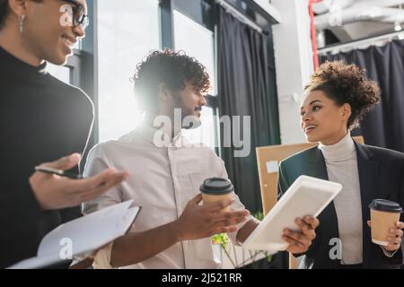 Fröhliche, multiethnische Geschäftsleute, die unterwegs Kaffee trinken und ein digitales Tablet im Büro unterhalten Stockfoto