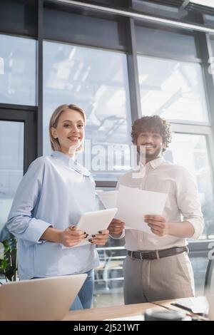 Lächelnde, multiethnische Geschäftsleute, die digitale Tablets und Dokumente im Büro halten Stockfoto