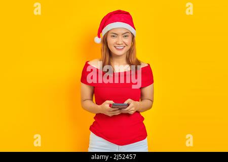 Porträt einer fröhlichen jungen Frau, die Weihnachten mit dem Smartphone auf gelbem Hintergrund trägt Stockfoto