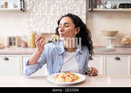 Junge lateinische Frau, die köstliche Pasta isst, leckeres hausgemachtes Mittagessen mit geschlossenen Augen genießt, in der Küche sitzt Stockfoto