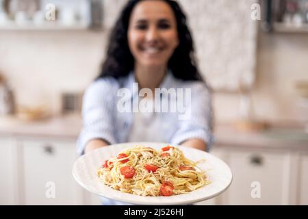 Frau zeigt hausgemachte Pasta im Gericht, dehnt Mahlzeit zur Kamera und lächelt, konzentrieren sich auf Teller mit Spaghetti Stockfoto