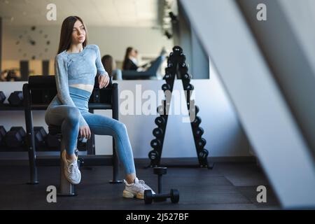 Nachdenkliches Mädchen sitzt auf einer Bank in einem Fitnessclub und ruht sich nach Übungen mit Hanteln aus Stockfoto