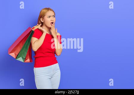 Überrascht junge asiatische Frau hält Einkaufstaschen mit offenem Mund und Blick auf Kopie Raum isoliert über lila Hintergrund Stockfoto