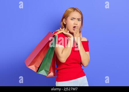 Überrascht junge asiatische Frau hält Einkaufstaschen mit offenem Mund und Blick auf Kamera isoliert über lila Hintergrund Stockfoto