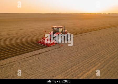 PRAG , TSCHECHISCHE REPUBLIK - MÄRZ 18 2022: Landwirtschaftliche Maschine bereitet lange Betten für die Pflanzung von Kartoffeln auf Kulturfeld. Der rote Traktor zieht den Pflug hinter die Luftaufnahme Stockfoto