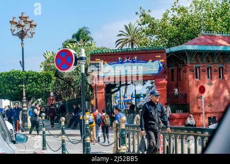 Sofia-Park-Eintrag mit einem Namensschild in arabischen Zeichen und französischer Sprache. Eine Menge Leute, die gehen und sitzen, Palmen und Lichtmast. Stockfoto