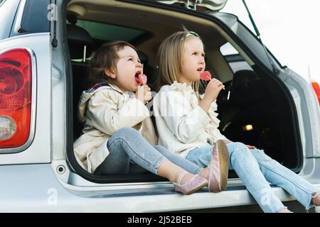 Zwei kleine Mädchen sitzen im Kofferraum eines Autos und essen Lutscher vor einer Autofahrt Stockfoto