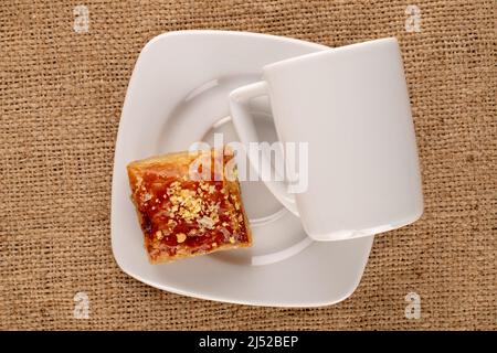 Süßer Baklava-Klassiker mit weißer Untertasse und Tasse auf Jute-Stoff, Makro, Draufsicht. Stockfoto