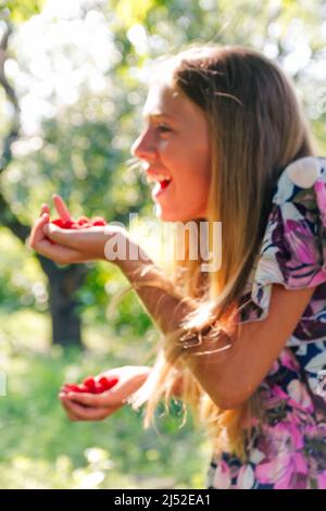 Defocus schöne lächelnde Teenager-Mädchen im Kleid lächelt vor grünen Sommer Hintergrund. Abstrakter Sommerhintergrund. Teen Mädchen hält Himbeeren Stockfoto