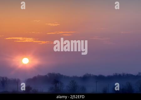 Wunderschöner Sonnenaufgang über der ländlichen Landschaft mit kalten nebligen Bäumen und orangefarbenem Himmel Stockfoto
