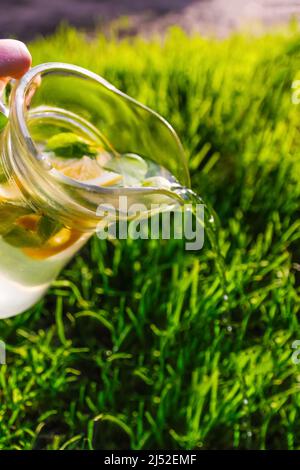 Unschärfe-Hand hält Glaskrug Limonade mit Zitrone und Minze auf natürlichem grünen Hintergrund. Krug mit einem kühlen Sommercocktail oder Punsch im Freien Stockfoto