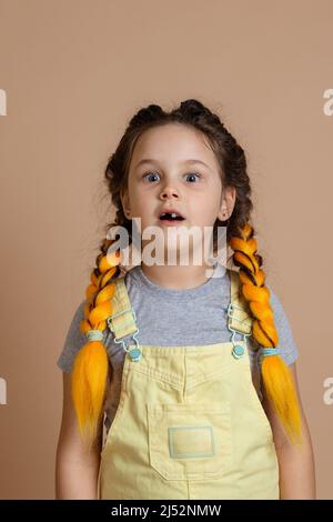 Schockiert schöne kleine Weibchen mit gelben Kanekalon-Pigtails, Blick auf die Kamera mit geöffnetem Mund mit fehlenden Zahn tragen gelben Jumpsuit und grau t Stockfoto