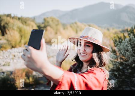 Schöne und lächelnde junge Reisende Frau, die einen Videoanruf von ihrem Handy in einem natürlichen Raum macht. Stockfoto