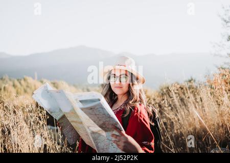 Junge Frau, die einen Hut, einen roten Poncho und einen Rucksack trägt, überprüft die Karte während der Wanderung, um die Natur zu erkunden. Stockfoto