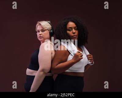 Zwei weibliche Freundinnen im Sport-BH stehen zusammen auf braunem Hintergrund. Frauen in Sportbekleidung stehen nach dem Training. Stockfoto
