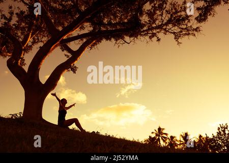 Junge Frau sitzt unter einem Baum mit ausgestreckten Armen und fühlt sich frei, und glücklich im Freien in einem wunderschönen Sonnenuntergang in der Natur Stockfoto