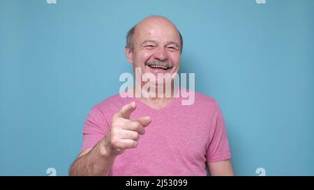 Älterer hispanischer Mann, der Sie anlacht und mit dem Finger auf die Kamera zeigt. Studio auf blauer Wand Stockfoto