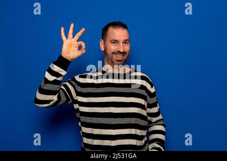 Ein spanischer bärtiger Mann in seinen 40s Jahren trägt einen gestreiften Pullover, der die OK-Geste macht, während er lächelt, isoliert über einem blauen Studiohintergrund Stockfoto