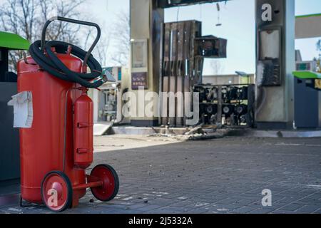 Roter Feuerlöscher auf dem Parkplatz nach dem Brand. Tankstelle mit einer kaputten Station durch eine Granatexplosion. Explodierte Tankstelle während des Krieges Stockfoto