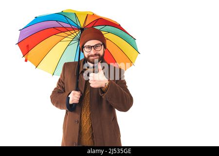 Bärtiger Mann, der bei Regen die Daumen hochstreckte. Hipster mit buntem Regenschirm. Rain man. Der farbenfrohe Regenschirm fällt immer ins Auge. Hochwertige Fotos Stockfoto