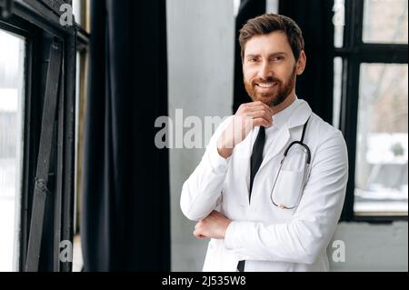 Porträt eines stolzen selbstbewussten kaukasischen professionellen Therapeuten, Chirurgen, Kardiologen in einer medizinischen Uniform und einem Stethoskop, der in einem Krankenhaus steht, auf die Kamera schaut und freundlich lächelt Stockfoto