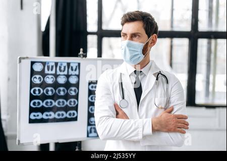 Nachdenklicher kluger kaukasischer Arzt, Allgemeinmediziner, Chirurg, Pneumologe, Brillenträger, Medizinische Uniform und Schutzmaske, stehen in der Klinik, Blick zur Seite mit gekreuzten Armen Stockfoto