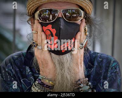 Älterer kaukasischer Mann mit verspiegelter dunkler Sonnenbrille und handbemalter schwarzer Stoffmaske (Gear of war) legt seine beringten Hände auf seine Wangen. Stockfoto