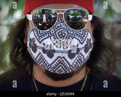 Der junge kaukasische Mann mit langen Haaren bedeckt sein Gesicht mit einer Gesichtsmaske aus nichtmedizinischem Tuch und trägt während der Pandemie eine verspiegelte tropfenförmige Aviator-Sonnenbrille. Stockfoto