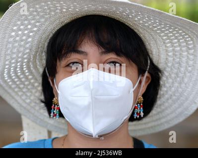 Die junge hübsche Mexikanerin mit bob-Haarschnitt, Ohrringen und braunen Augen trägt während der globalen Coronavirus-Pandemie einen weißen Sonnenhut und eine weiße KN 95 Gesichtsmaske. Stockfoto