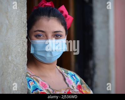 Positive junge mexikanische Frau mit lächelnden Augen und blauem Augen-Make-up trägt ein farbenfrohes traditionelles Yucatan Maya-Kleid und eine hellblaue OP-Gesichtsmaske. Stockfoto