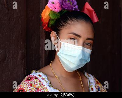 Die attraktive junge mexikanische Frau mit Blumen in gesägtem Haar trägt ein farbenfrohes, traditionelles Yucatan Maya Huipil-Kleid und eine chirurgische Gesichtsmaske. Stockfoto