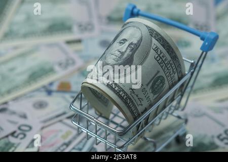 In einen Schlauch gerollte Dollars liegen in einem Einkaufskorb. Der Korb steht auf einem verschwommenen Geldhintergrund. Stockfoto