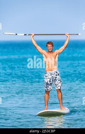 Glücklicher Wassersportler mit Spaß auf Stand-up-Paddleboard sup halten Paddel in der Luft in Erfolg auf Paddle-Board-Rennen. Gut aussehender, junger Athlet Stockfoto