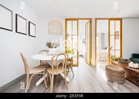 Modern eingerichtetes Wohnzimmer mit einem runden weißen Esstisch aus Holz, Korbsitz, Holzstühlen und Buntglasfenstern mit gelbem Metall Stockfoto