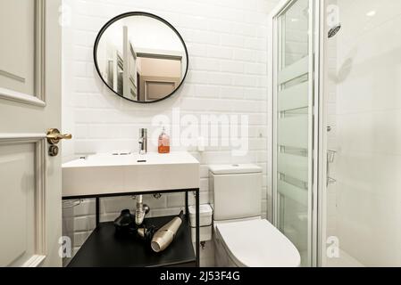 Badezimmer mit Duschkabine mit Glasschiebeschirm, runder Spiegel mit schwarzem Rahmen, Waschbecken aus weißem Porzellan auf schwarzem Metallschrank Stockfoto