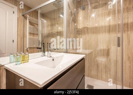 Badezimmer mit quadratischem rahmenlosem Spiegel, weißem Porzellanwaschbecken auf Holzschrank, weißem Handtuchhalter, begehbarer Dusche mit Bildschirm und cremefarbener Marmorverkleidung Stockfoto