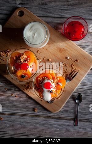 Zwei Glasbecher mit Aprikosenscheiben, Müsli-Joghurt und Kirschen auf der Oberseite, mit Besteck an der Seite und auf einem Schneidebrett, über einem Holztisch. Stockfoto