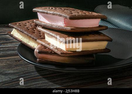 Eiscreme-Sandwiches auf einem schwarzen Teller mit einer blauen Serviette unten über einem Holztisch mit schwarzem Hintergrund, beleuchtet durch eine Fensterbeleuchtung Stockfoto