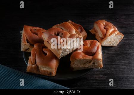 Dunkle Food-Fotografie, Zimtrollen mit Rosinen auf einem schwarzen Teller mit blauer Serviette über einem rustikalen Tisch. Chiaroscuro-Food-Fotografie Stockfoto