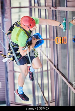 Arbeiter beim Bergsteigen waschen Glasfenster des Hochhauses, hängen an einem Kletterseil. Man Fensterputzer in Schutzhelm Reinigung Wolkenkratzer Fassade. Draufsicht. Stockfoto