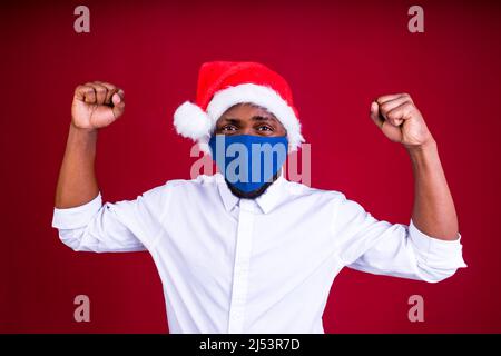 afroamerikanischer Mann in santas Hut trägt Maske im roten Studio Hintergrund Stockfoto