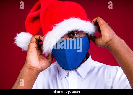 afroamerikanischer Mann in santas Hut trägt Maske im roten Studio Hintergrund Stockfoto