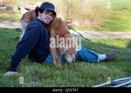 Ein hübscher Kaukasusmann sitzt allein auf einem grasbewachsenen Hügel mit Naturhintergrund und ein liebevoller goldener Retriever-Hund auf seinem Schoß mit einem Paar Krücken auf dem Schoß Stockfoto