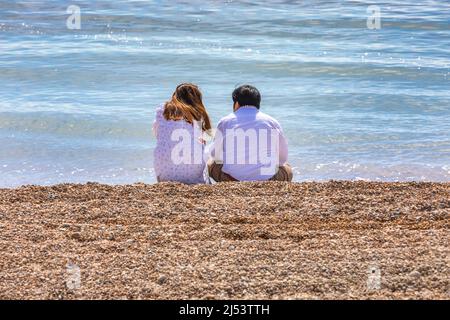 Ein junges Paar, das an einem Kiesstrand an einem blauen Meer sitzt. Stockfoto