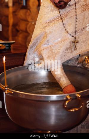 Nicht erkennbarer bärtiger Priester in silberner Soutane steht am Metallbecken und weiht Wasser für die Zeremonie der Säuglingstaufe. Stockfoto