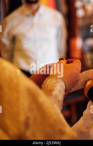 Zugeschnittenes Foto des Priesters in zeremonieller Soutane halten Flasche mit heiligem Öl für die Salbung Kind während der Zeremonie der Taufe. Stockfoto