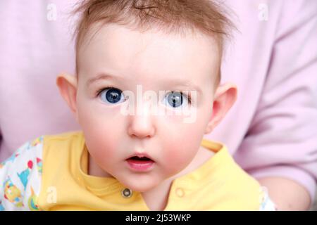 Überraschte den kleinen Jungen mit großen blauen Augen und zerzausten Haaren in den Armen seiner Mutter. Porträt eines verängstigten Kindes mit einer unordentlichen Frisur Stockfoto